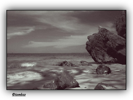 Black-and-white seaside landscape featuring a rocky shoreline. In the foreground, several large, irregularly shaped rocks protrude from the water, their rough, jagged texture visible. The sea is calm, with gentle waves softly blending into the shore. Sunlight reflections create shimmering streaks on the water’s surface, enhancing the contrast between the light and dark areas of the image. In the background, the open expanse of the sea meets the horizon, where a faint coastline adds depth to the composition. The sky is covered in soft, elongated clouds, forming a dynamic yet tranquil backdrop for the entire scene.