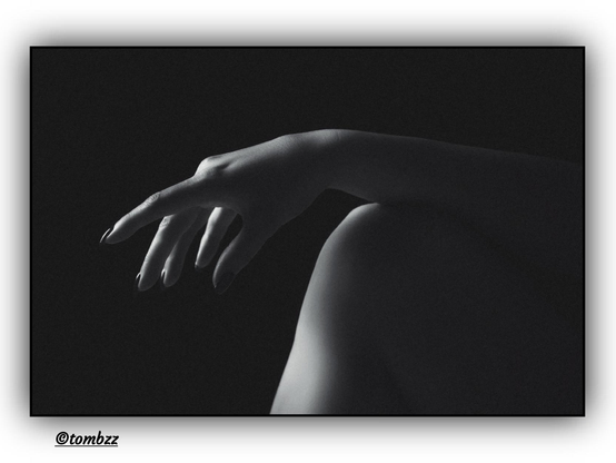 The photo captures an elegant, black-and-white composition focusing on a delicate detail of a woman's silhouette. At the center of the image, a slender leg provides support for a gracefully positioned hand. The woman's fingers form a sensual gesture, subtly emphasizing the softness and fluidity of movement. The contrast of the dark background enhances the mystery of the scene, bringing out the subtle interplay of light and shadow, which gives the composition a deep, elegant character. The play of light and shadow adds depth to the image, creating a magical, refined atmosphere.