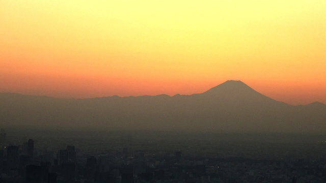 The shot is against the setting sun backlighting the famous mountain on the right. Other mountains form a ridge to the left of it. The cityscape of Tokyo is barely discernable in a sea of barely lit air pollution.