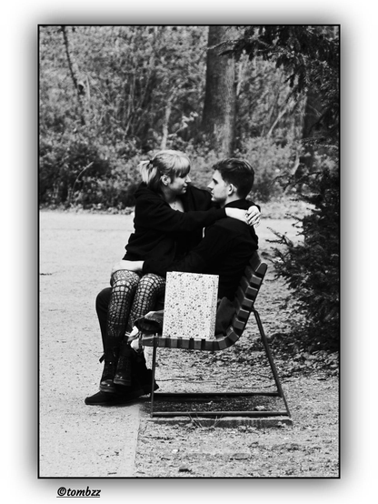 The photo depicts a couple in a park. The young man is sitting on a bench, while the young woman is on his lap, their closeness indicating a romantic bond. She is wearing fishnet stockings, a short skirt, and chunky platform shoes, with her long hair cascading freely over her shoulders. The young man is dressed in dark pants, a t-shirt, and an open jacket. A paper gift bag sits on the bench beside them, suggesting a special occasion. In the background, trees and bushes add to the intimate atmosphere. The photograph is in black and white, emphasizing its nostalgic and emotional tone.