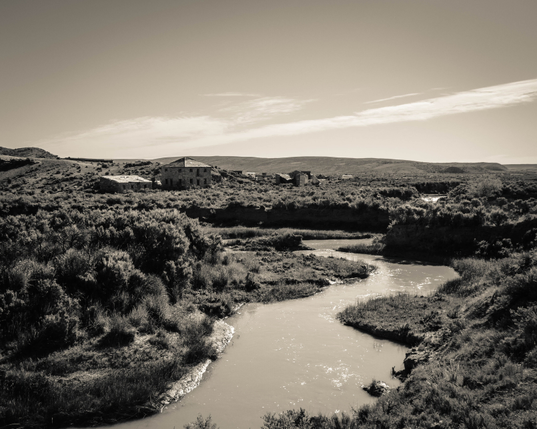 A sepia toned photograph of an abandoned farm in Wyoming.  A stream meanders in the foreground, flanked by sagebrush covered fields.  The farm house and its buildings, clustered close by, are in various stages of decay.