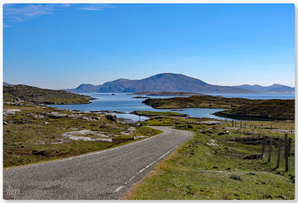 Eine einspurige Straße führt auf eine Meeresbucht zu. Das Wasser ist blau, genau wieder der wolkenlose Himmel. Im Hintergrund Berge. Rechts und links der Straße Felsen, Steine und etwas Grün.

A single-track road leads to a bay. The water is blue, just like the cloudless sky. Mountains in the background. Rocks, stones and some greenery to the right and left of the road.