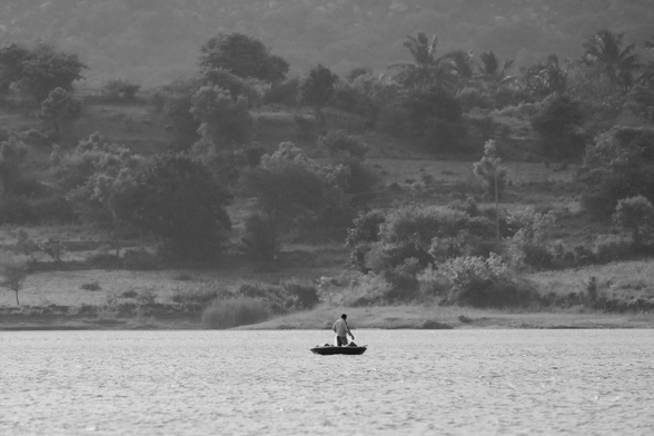 A lone fisherman on a small coracle on a wide lake, with tree-covered hills rising in the background, in black and white.
