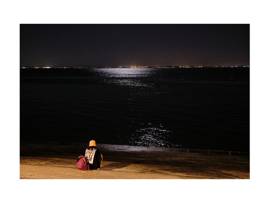 lonesome person from the back sitting at the riverside of tejo river in lisbon at night, staring over the water, full moon shining from above, scattered reflexions in black water