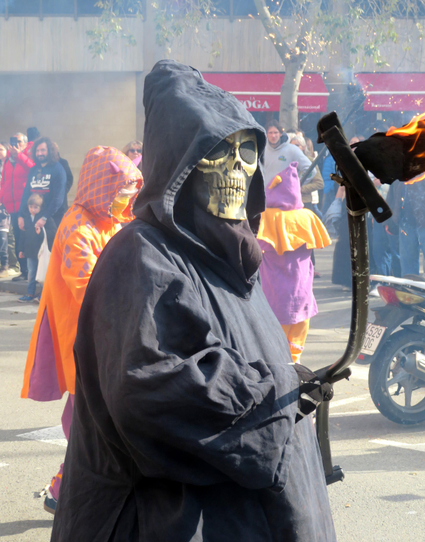 The image depicts a street scene with a person in the foreground wearing a black hooded robe and a skull mask with eye holes, holding a scythe with a flame at the end. The person is standing on a street with a crowd of people in the background, some of whom are wearing colorful costumes, including an orange and purple outfit. The background shows a building with a red awning and a tree, with smoke visible in the air, suggesting a festive or celebratory event. The overall atmosphere appears to be lively and communal, with people gathered for a public event.

Provided by @altbot, generated privately and locally using Ovis2-8B

🌱 Energy used: 0.172 Wh