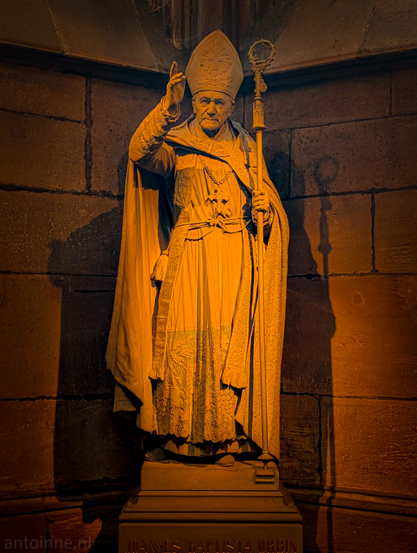 A large, light-coloured stone statue of a religious figure is standing within an old stone building. The statue wears a tall, pointed miter on his head. A cross hangs around its neck. His right arm is raised with his hand open, as if blessing or addressing someone. Johann Baptist Orbin was the Archbishop of Freiburg.