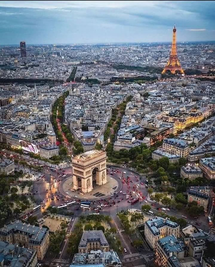 The image depicts an aerial view of a cityscape featuring prominent landmarks. At the center is the Arc de Triomphe, surrounded by a busy roundabout with multiple roads radiating outwards. To the right of the Arc de Triomphe stands the Eiffel Tower, illuminated and towering over the city. The city is densely packed with buildings, and there are visible green spaces and avenues leading towards the horizon. The overall scene suggests it is Paris, France, known for these iconic structures.