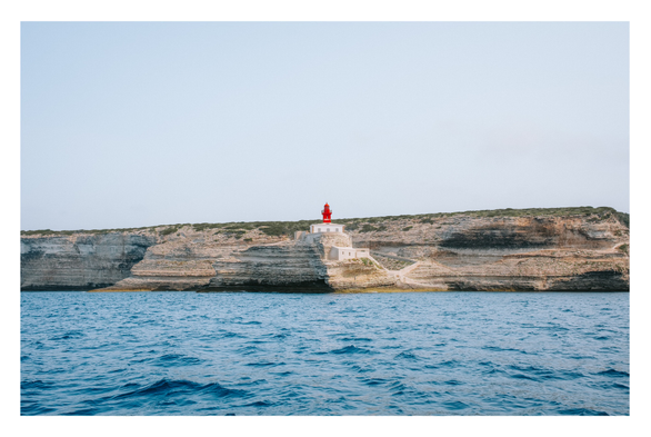 Photo of a red Lighthouse in the distance standing atop of white cliffs. Water in the foreground. Light blue sky.