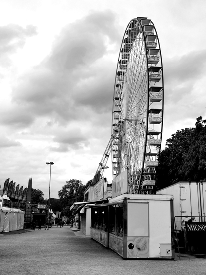 Vue en noir et blanc d'une foire attractive peu fréquentée. Au premier plan se dresse la grande roue sur fond de ciel chargé