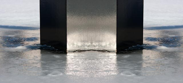 In a frozen pond, in the middle, stands a broad, black column, which reaches from the center of the photo to the top. The sun reflects in the shiny column. The ice is wet and the object reflects in it. About two-thirds of the way through the photo, the ice is dry and white snow-covered.