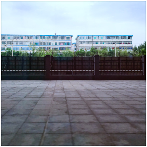 A deserted platform in the foreground; a fence, tracks, and greenery in the center; and flat buildings in the background.