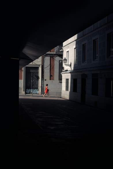 Una fotografia en la que una mujer de rojo pasea por la calle junto a una niña justo por la zona iluminada entre edificios que hacen de marco oscuro