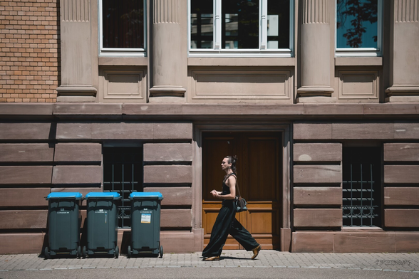 The image shows a street scene with a single person walking briskly from left to right along a paved sidewalk. The man is dressed in a sleeveless black outfit and brown shoes, carrying a black bag over his shoulder. He walks in front of a large stone building with neoclassical architectural details, including columns and large windows above a series of grated basement windows. The door behind her is wooden with a darker wood finish.

To the left of the person, there are three large dark-colored garbage bins with blue lids positioned on the sidewalk. The wall behind them transitions from a reddish-brown stone facade to a section of tan brickwork.

The image looks to have signs of pincushion distortion (a type of lens distortion where straight lines bow inward). But in fact it does not have any distortion. It is due to the  curved appearance of the building’s left wall and the sidewalk at the bottom of the frame.