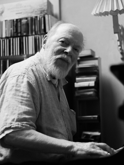 Black and white photo of an old man playing the piano. He looks in the camera with a little smile. 