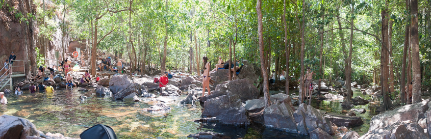 Looking downstream from the outflow from a waterfall plunge-pool. The watercourse is very wide here, yet to gather back into a stream, and shallow water runs around numerous rocky outcrops and even trees growing in the creek bed. Downstream is quite heavily timbered,  and dappled green light filtering through all the foliage illuminates the scene. The spot is very popular with both tourists and locals, and there are a lot of people in swimwear scattered through the scene.