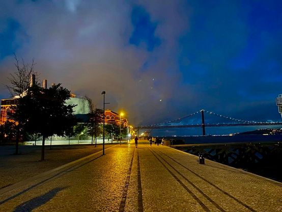 A scenic nighttime view of a waterfront promenade featuring a bridge illuminated by lights. Nearby buildings, partially obscured by trees, are softly lit, and a pathway lined with cobblestones leads toward the water. The sky is cloudy with hints of color,