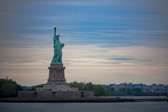 Statue of Liberty against a cloudy sky with waterfront and city skyline in the background.