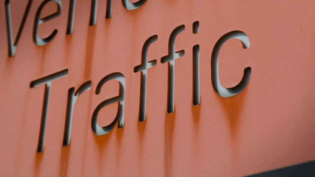 Close up of a sign that uses an orange sheet of metal with the letters cutout in front of a gray sheet of metal. Reads “Traffic” with a cutoff word above. Shadows and discoloration make the letters look neat.