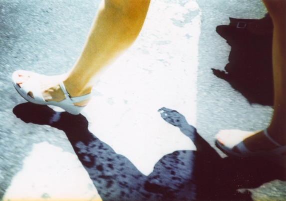 Detail of feet and shadow of a woman wearing white sandals while on a zebra crossing under the sun. Taken on Agfa RSXII analogue film using a Cosina CX2 camera.