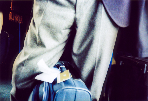 Detail of a man legs wearing grey pants keeping a blue travel suitcase with tags between his legs. Taken on Agfa CT Precisa analogue film using a Cosina CX2 camera.