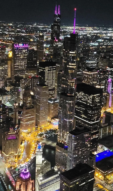 The image depicts a nighttime cityscape with numerous illuminated skyscrapers. The buildings are densely packed, and their windows glow with various colors, including purple and pink lights on some of the taller structures. A river winds through the city, reflecting the lights from the surrounding buildings. The city appears to be bustling with activity, showcasing a vibrant urban environment. This cityscape is likely Chicago, identifiable by the distinctive architecture and the river.