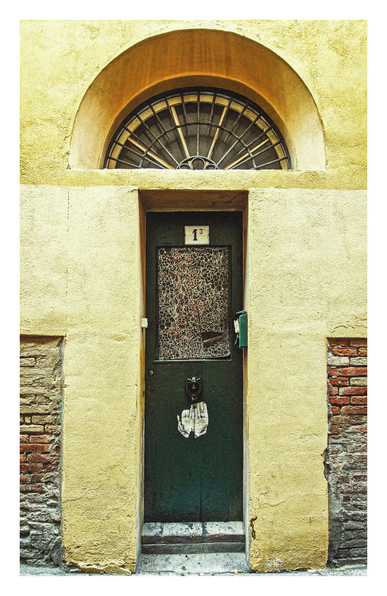 The photo is a close-up view of a doorway set into a pale yellow stucco wall. The door is dark green colour with the top half that has a glass pane. Through the glass pane there is some floral paper covering the glass from the inside. Under the glass is a metal door knocker in the shape of a horse head surrounded by a horse shoe. There is a old weathered sticker on the door of open hands underneath the glass panel.and the door knocker. Above the door is a half-circle, segmented window set into the wall.