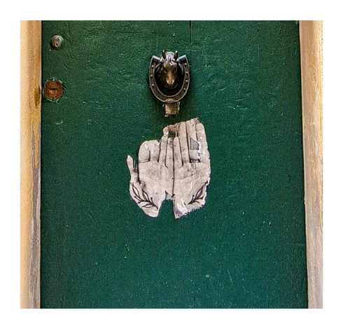 This photo is a close up of the dark green door with a horseshoe-shaped door knocker featuring a horse's head. Below the knocker is a partially torn sticker depicting two hands with leaves at the base of the palms, which appear palm forward to the viewer. The sticker is a white colour.