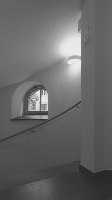 View of a semicircular window to the outside (windows on another building are seen) above a spiral staircase. Surfaces with hardly any structure and the black-and-white editing emphasize the gemoetries of the different lines.
