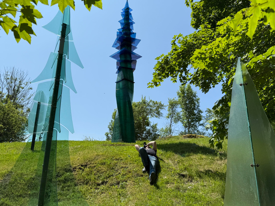 Some guy lies on his back on a grassy slope beneath a tall blue glass sculpture, pointing his camera upwards