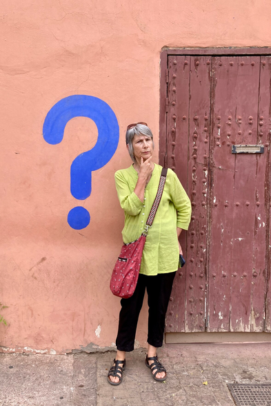 Lady standing in front of wall painted pinkish, that has a large blue question mark painted on it. A reddish door is on her other side.