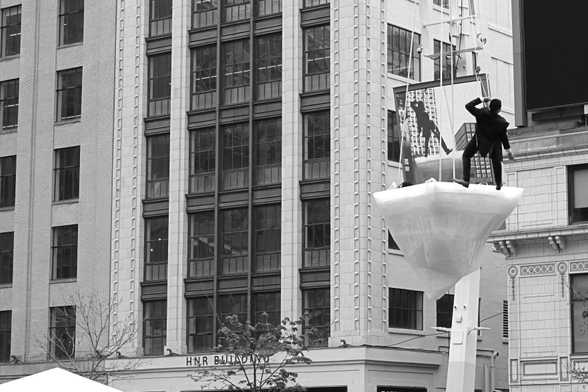 In the upper right of the frame, as part of the Luminato festival, a man stands on an artificial iceberg suspended from a crane above the square as part of a performance piece. Behind him is his image on a huge video screen, showing him from a different angle. The upper left of the frame is filled with the facade and windows of the office building beyond. 