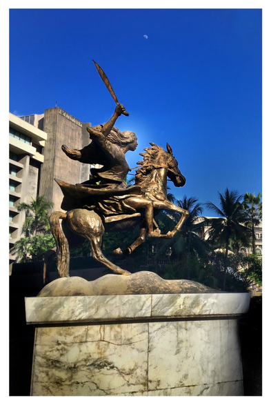 This is a monument to the Filipina revolutionary Gabriela Silang, which was designed by Jose M. Mendoza and stands in the central business district of Makati, Metro Manila.

Gabriela Silang led her troops and the people of her province in a resistance movement against the Spanish colonial government after her husband was assassinated in 1763. She herself was eventually captured and executed, but others took up the cause — because the struggle for liberation is not the work of one person or one lifetime. May we share that spirit today as we do what we can, in actions big and small, to dismantle systems of oppression until everyone receives justice and dignity, until everyone is free.

Image description: A bronze statue of a young woman, facing right and riding a horse that rears up with its forelegs raised. The woman’s hair flows back as if wind-blown and she wields a raised bolo in her right hand. The statue stands on a wide marbled pedestal. Behind it is a line of palm trees and a high-rise building with the words THE PENINSULA at the top of its facade. High overhead in a blue sky, a tiny moon looks down on the scene.
