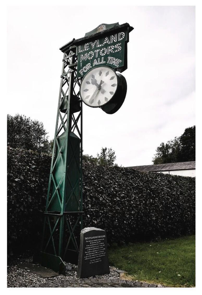 Colour portrait photograph showing a clock hanging from a green gantry tower.  Above the clock are the words Leyland Motors For All Time.