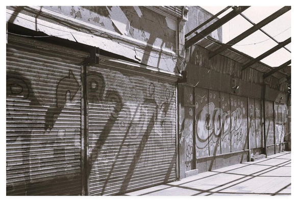 Black and white film photograph showing graffiti on the shuttered properties of an abandoned shopping arcade.