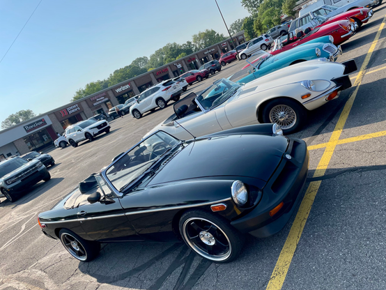 Photograph tilted down and to the left of a black rubber-bumper 1976 MGB next to a white 1974 Jaguar XKE (aka E-Type) in a strip mall parking lot. Several other small British cars can be seen behind them.
