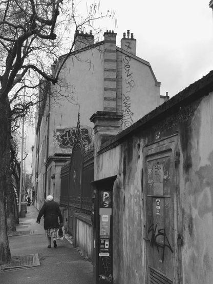 Dans une rue en noir et blanc, devant une grille passe, une femme portant un cabas chargé. Le pignon d'un immeuble voisin domine la scène.
