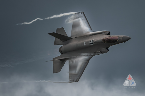 Un avion de chasse furtif de type Lockheed Martin F-35 effectue un virage à grande vitesse dans le ciel, produisant des traînées de condensation sur ses ailes. L’appareil est vu en vol incliné, avec la verrière du cockpit bien visible et les détails de sa structure aérodynamique mis en valeur par l’éclairage et les effets de condensation. Le logo « SKYWORX » apparaît en bas à droite de l’image.