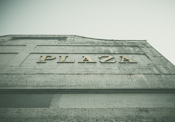 A weathered brick building facade photographed from a low angle against an overcast sky. The word "PLAZA" is prominently displayed in raised block letters across the upper portion of the wall, set within a recessed rectangular panel with decorative molding. The brick wall shows signs of age with some staining and discoloration, particularly visible around the lettering area. The building's architectural details include stepped horizontal bands and trim work typical of mid-20th century commercial construction. The photograph is taken from below, emphasizing the building's height and giving the lettering a bold, imposing appearance against the pale gray sky.