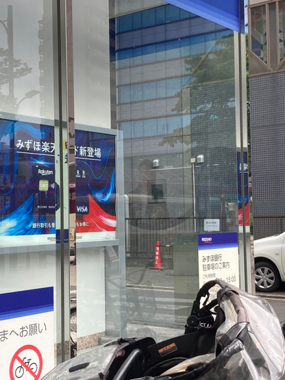 A street scene featuring a glass-encased bank entrance with promotional signage for Mizuho and Rakuten. A stroller is positioned in the foreground, and a traffic cone is visible in the background, along with several buildings reflecting in the glass.