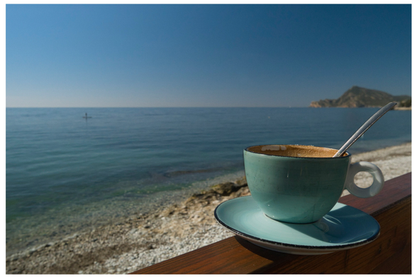 auf einer Holzbalustrade einer Imbissbude,  un chiringuito, steht eine blaue Kaffeetasse mit Unterteller in der ein silberfarbener Löffel steckt. Der braune angetrocknete Milchschaum ist sichtbar. Dahinter Kieselstrand und das Meer, wolkenloser Himmel.