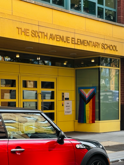 Color photo, in the lower left-hand corner is a small bright red car parked in front of a building that says Sixth Avenue Elementary School. The building is painted a bright yellow and on a glass window to the side hangs a large pride flag