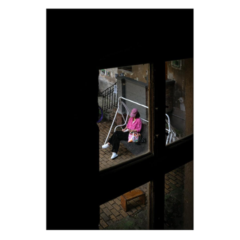 The picture was taken from the window of the stairwell of an old apartment building in the city center. In the courtyard, a woman wearing a pink top was sitting on a white swing bed and writing in a notebook. On the left side of the picture, you can see the darkness of the stairwell and the frame of the window through which the photo was taken.