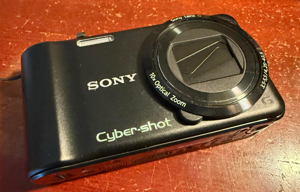 A Sony Cyber-shot digital camera resting on a wooden surface. The camera features a prominent lens with a 10x optical zoom.