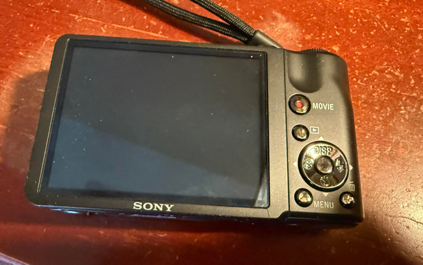 A Sony digital camera with a blank screen and various control buttons, resting on a wooden surface.