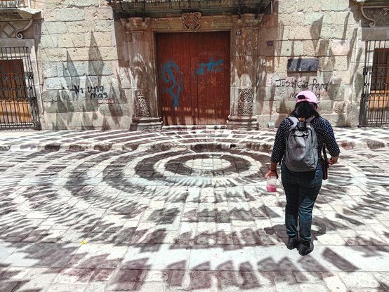 A Zapotec woman wearing a pink ball cap, black jeans, and a black jacket, carrying a black backpack, stands on a cobblestone street facing away from the viewer, towards a stone building with a wooden door and two barred windows. Both the walls and door have grafitti on them. There is a shadow design of broken concentric circles on the street and the lower part of the building, cast by decorative paper flags hung above out of frame.