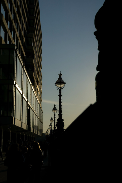 On the left a building with many stories, the first two all large square glass fronted, frames shining in bright low sunlight, the remainder further back, recessed in concrete, all the windows reflecting the faded blue sky in the centre. The right-hand edge is all the silhouette of what looks to be the curving and moulded base of an ornate lamppost atop a wall, going by the examples stretching off into the distance. At the bottom, in the deep shadow of the wall, hints of people walking and catching the light.
