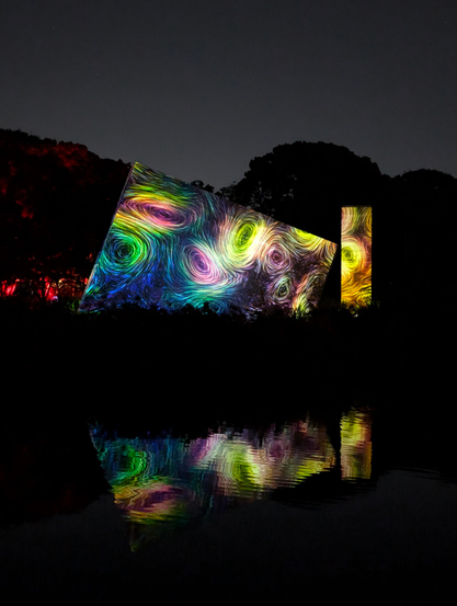 At night, a tilted rectangular block can be seen in the middle of the photo. Colorful vortex-like shapes, that remind one of a photo of galaxies in space are projected onto it. The water of the lake in front of the block reflects the block.