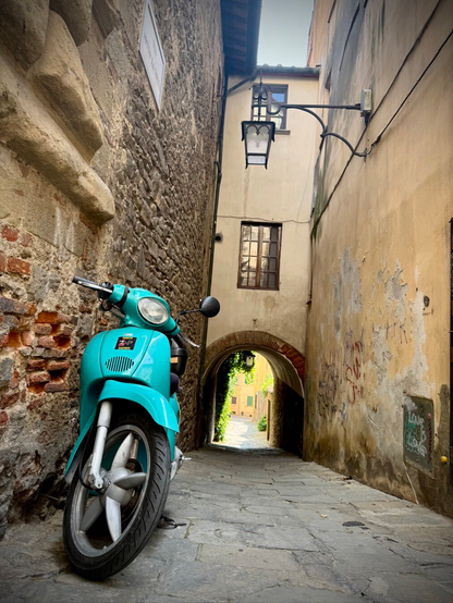 A vintage turquoise scooter is leaned against a stone wall in a narrow alleyway. The alley features a mix of textured stone and plaster walls, a hanging lantern, and a view of an archway leading to a vibrant, green-lined area in the background