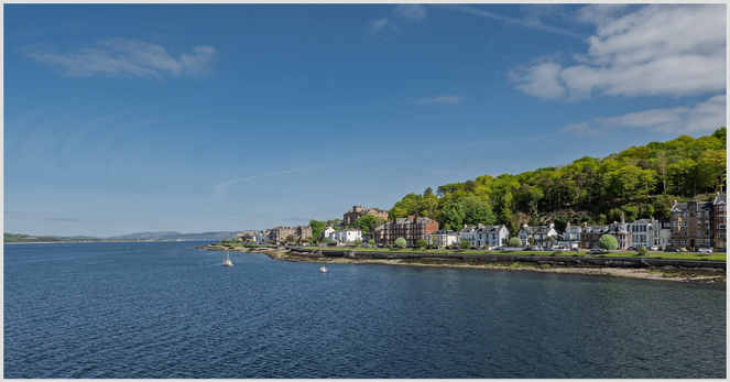 A coastal view featuring a calm blue sea with a few small boats, flanked by a shoreline of houses and buildings. In the background, there's a steep, green hillside. The sky is clear with a few clouds.