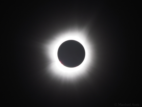 Photo of a total solar eclipse. Black sky, black moon and a bright corona with some glowing red spots along the rim.